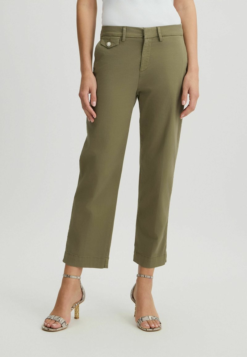 Femme portant un pantalon court vert olive et des sandales à talons hauts imprimé serpent, debout devant un fond uni.