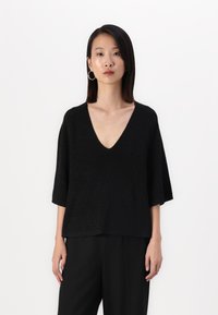 Top de malha preto com decote em V e mangas curtas, apresentando uma textura e brilho subtil. Combinado com calças pretas largas. Design limpo e moderno.