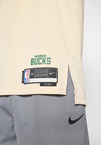 Krémový basketbalový dres s nápisom "MILWAUKEE BUCKS" v zelenej farbe, logo Nike a štítok s veľkosťou. Obsahuje sieťovanú látku a bočné rozparky pre ventiláciu.