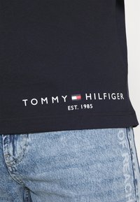 Granatowa bawełniana koszulka z białym napisem "TOMMY HILFIGER EST. 1985" na dolnym brzegu, noszona z jasnoniebieskimi dżinsami.