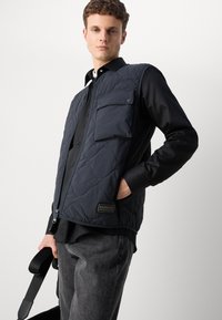 Vest cu umplutură de culoare bleumarin, cu fermoar frontal, un buzunar pe piept și un patch cu logo. Purtați peste o cămașă neagră și asociați cu pantaloni gri.