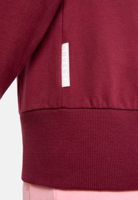 Bordeauxroter Pullover mit geripptem Saum und Bündchen. Verfügt über ein weißes Stofflabel an der Seite. Glatte Textur, die ein lässiges Design zeigt.