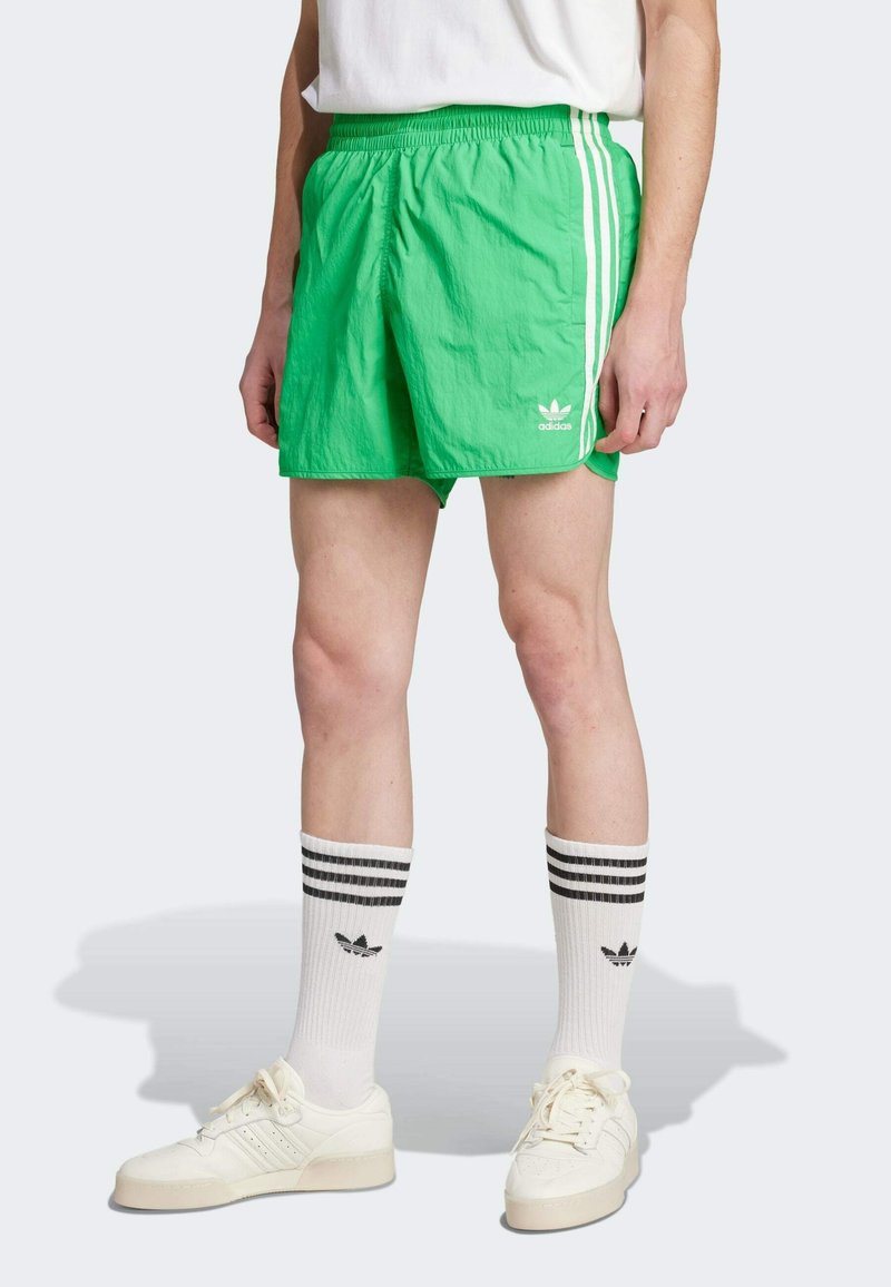 Πράσινο νάιλον σορτσάκι με λευκές ρίγες στο πλάι και λογότυπο Adidas. Φοριέται με λευκές κάλτσες που έχουν μαύρες ρίγες και λογότυπο Adidas, συνδυασμένο με λευκά παπούτσια.