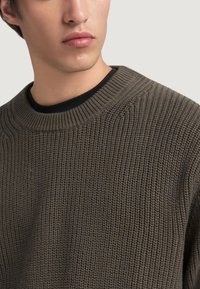 Jersey de punto en verde oliva oscuro con una tela texturada, cuello redondo y acentos de canalé. Camiseta negra visible en el escote.