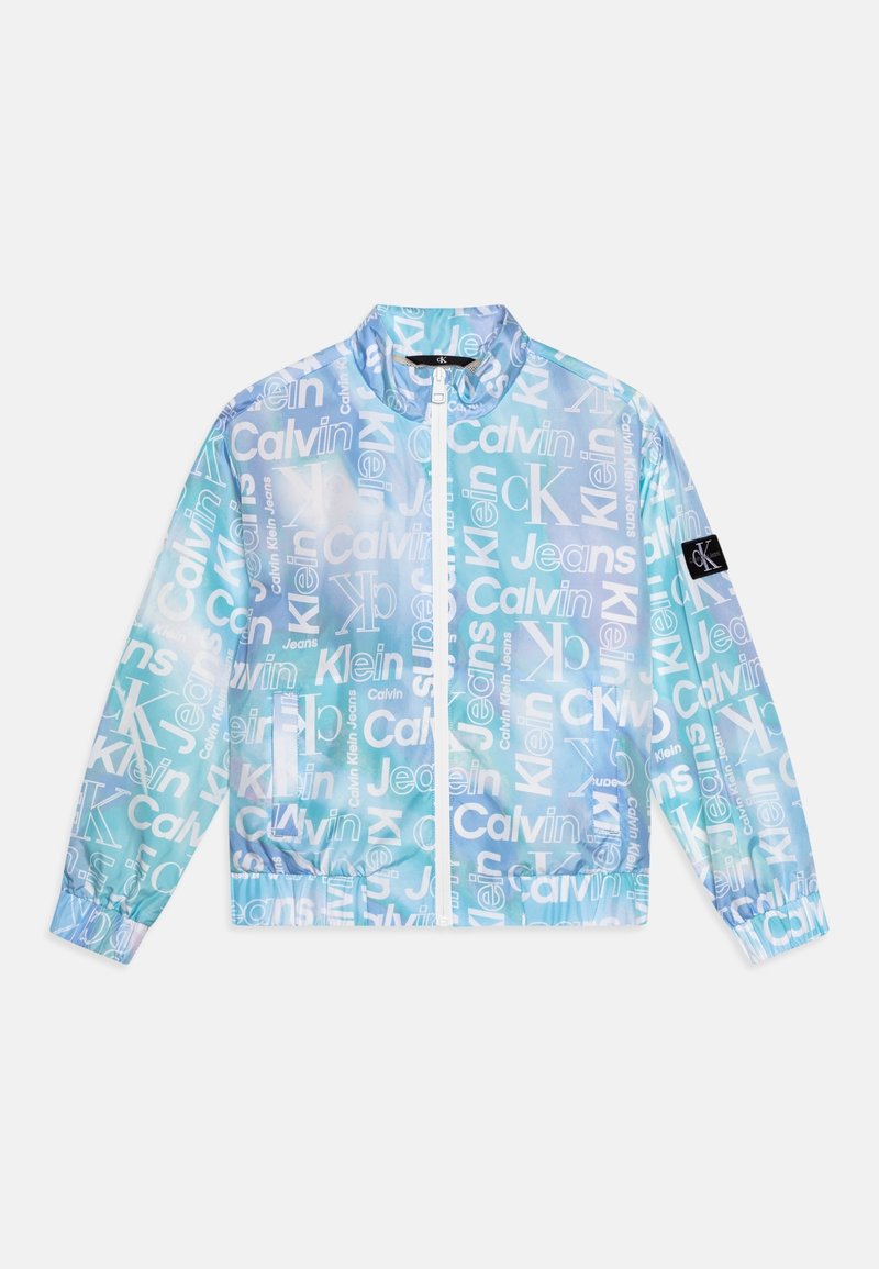 Coupe-vent bleu translucide avec des logos blancs Calvin Klein. Il comporte une fermeture éclair à l'avant, des poignets élastiques et un logo en patch sur la manche. Texture lisse.