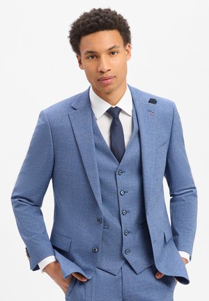 Mann mit einem hellblauen Dreiteiler-Anzug, weißem Hemd und dunkelblauer Krawatte, der mit den Händen in den Taschen vor weißem Hintergrund posiert.