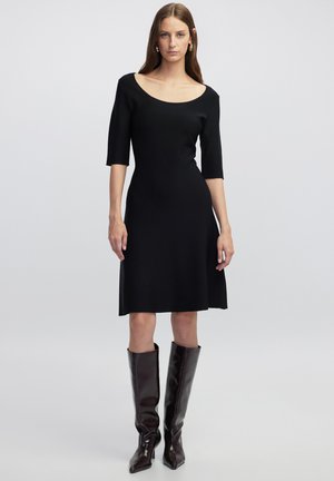 Robe en maille noire avec un décolleté dénudé, des manches trois-quarts et une jupe évasée, associée à des bottes marron brillantes jusqu'aux genoux.