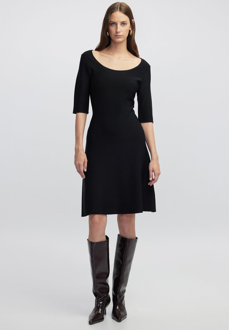 Zwarte gebreide jurk met een off-shoulder halslijn, driekwart mouwen en een uitlopende rok, gecombineerd met bruin glanzende laarzen tot de knie.
