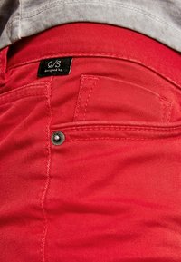Vaqueros de mezclilla rojos con un acabado texturizado, que presentan un pequeño bolsillo delantero y una etiqueta negra "Q/S diseñado por" en la cinturilla.