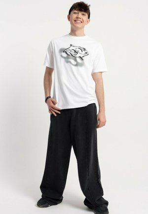 Nastolatek uśmiechający się, ubrany w białą koszulkę z abstrakcyjnym czarnym wzorem, luźne czarne spodnie i czarne sneakersy, stojący na białym tle.