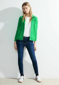 Felpa verde con zip, t-shirt bianca, jeans blu scuri e sneakers bianche. La felpa presenta un cordino e tasche frontali.