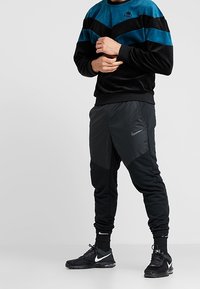 Svart och teal velourtröja med chevrondesign, i kombination med smala svarta joggers med Nike-logotyp och texturerade detaljer.