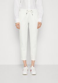 Bílé tapered jogger kalhoty s gumou v pase, bočními kapsami a hladkým materiálem. V kombinaci s bílými teniskami s červenými akcenty.