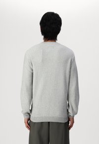 Pull en maille grise à rayures, à manches longues, avec un col rond et des poignets ajustés, arborant un motif texturé, porté sur un pantalon ample.
