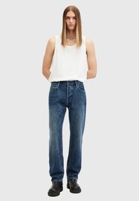 Jeans in denim, blu medio sbiadito, design a gamba dritta, abbinati a un top bianco senza maniche. La modella indossa stivaletti neri in pelle con tacco, mani ai fianchi.