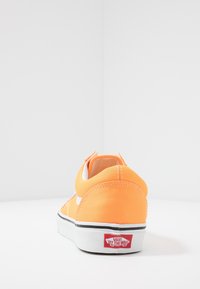 Oranžové tenisky Vans ukazující zadní část paty s bílou gumovou podrážkou a červenou visačkou "Vans Off The Wall" na bílé podložce.