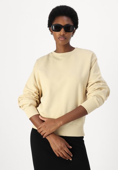 Σα sweatshirt σε κρέμα απόχρωση με λαιμόκοψη τύπου crew, ριμπ μανσέτες και άνετη εφαρμογή. Το μοντέλο φορά μαύρα γυαλιά ηλίου και στέκεται σε ένα απλό φόντο.