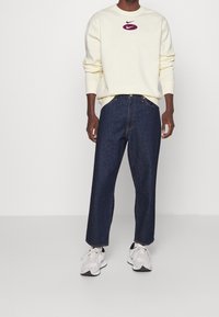 Bílá mikina s fialovým logem Nike, v kombinaci s volnými tmavě modrými džínami a světle šedými sportovními botami. Jednoduchý design, volný střih.