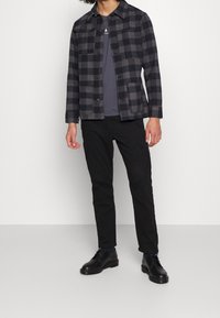 Veste à carreaux noir et gris en tissu, avec un devant à boutons, deux poches et un col, associée à un pantalon cargo noir et des chaussures.