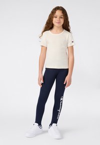 Kremowa koszulka z krótkim rękawem z logo na piersi, zestawiona z ciemnoniebieskimi legginsami z białym nadrukiem logo oraz białymi sneakersami.