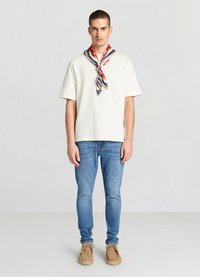 Chemise crème à manches courtes avec un col rond, associée à un jean skinny bleu clair et des chaussures en daim beige. Écharpe à motifs rouges, bleus et jaunes autour du cou.
