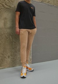Svart kortärmad Nike-t-shirt, beige slim-fit byxor och sneakers med orange detaljer. Väggen har en texturerad yta. Golvet är slätt och ljusgrått.