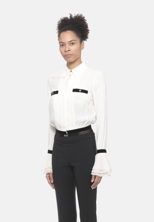 Donna che indossa una camicetta bianca con dettagli in velluto nero e polsini plissettati, abbinata a pantaloni neri a vita alta con una cintura.