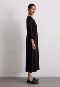 Czarna sukienka z długim rękawem i wcięciem w talii, wykonana z miękkiego materiału, opadająca do połowy łydki. Połączona z czarnymi loafersami, modelka stoi w profilu.