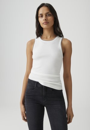 Vrouw draagt een wit mouwloos topje met zwarte afwerking en zwarte spijkerbroek tegen een effen lichtgrijze achtergrond.