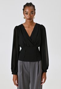 Blusa nera con design avvolgente, maniche a sbuffo in tessuto trasparente e vita aderente con doppi bottoni. Abbinata a pantaloni grigi.