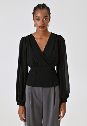 Blusa nera con design avvolgente, maniche a sbuffo in tessuto trasparente e vita aderente con doppi bottoni. Abbinata a pantaloni grigi.