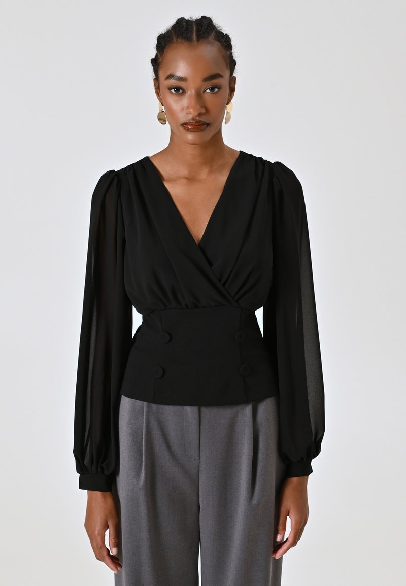 Blusa nera con design avvolgente, maniche a sbuffo in tessuto trasparente e vita aderente con doppi bottoni. Abbinata a pantaloni grigi.