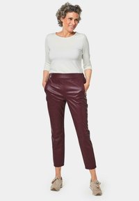 Bordeaux leren broek met voorzakken, gecombineerd met een witte, aangesloten lange mouw top en beige sneakers. Het model staat in een ontspannen houding.