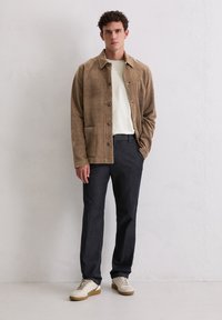 Tan suède jas met knoopsluiting, voorkantzak; gedragen over een wit shirt en donkere broek. Schoeisel: beige sneakers met bruine zolen.