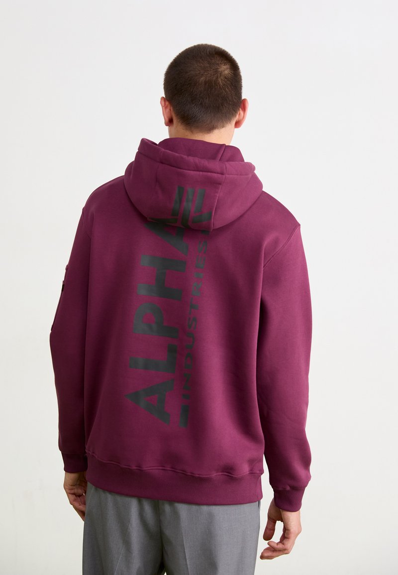 Burgunderfarget hettegenser med stor svart "ALPHA INDUSTRIES" logo printet vertikalt på ryggen. Myk stoff med romslig hette og ribbestrikkede ermer.