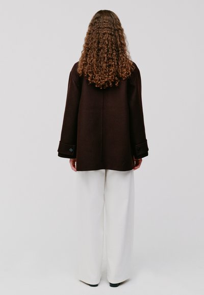 Persona con lunghi capelli ricci, che indossa un cappotto scuro e pantaloni bianchi larghi, in piedi davanti a uno sfondo bianco semplice.
