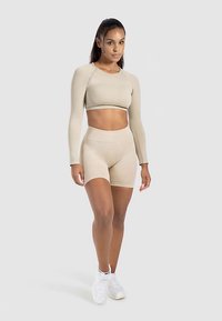 Beige crop top met lange mouwen gecombineerd met high-waisted shorts, gemaakt van rekbare stof. Model draagt witte sneakers, tegen een lichte achtergrond.