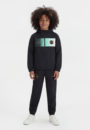 Enfant aux cheveux bouclés porte un survêtement Nike noir avec des panneaux turquoise et blancs, debout avec les mains détendues, baskets blanches, fond neutre.