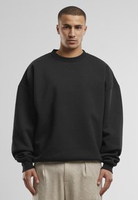 Felpa nera oversize con colletto rotondo e spalle scese, caratterizzata da una texture liscia e polsini a costine. Abbinate a pantaloni chiari.