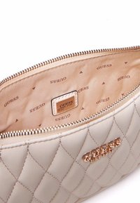 Pochette trapuntata color crema con logo "GUESS" in tonalità oro, chiusura con zip e interni beige con il nome del marchio stampato in un motivo ripetuto.