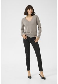 Maglione leggero marrone chiaro con scollo a V e maniche lunghe, con bottoni decorativi, abbinato a jeans skinny neri e tacchi neri, in piedi contro uno sfondo bianco.
