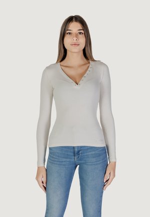 Top lungo a maniche con costine beige, con scollatura a V, decorato con piccoli bottoni e un sottile dettaglio con logo all'orlo. Abbinato a jeans blu.