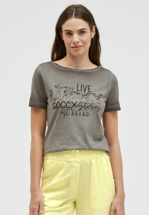 Young woman with long brown hair wearing a gray graphic t-shirt and light yellow pants, standing against a plain background.