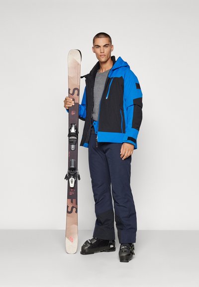 Skiausrüstung mit einer blauen und schwarzen wasserdichten Jacke, grauem Shirt, schwarzen Skihosen und schwarzen Skischuhen; hält braune Skier mit Bindungen.