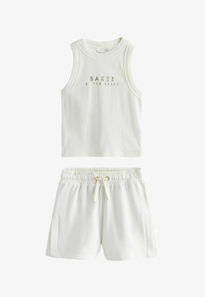 Jeune femme debout portant un haut blanc sans manches, un short blanc à cordon, des chaussettes blanches et des baskets blanches sur un sol et un mur gris.