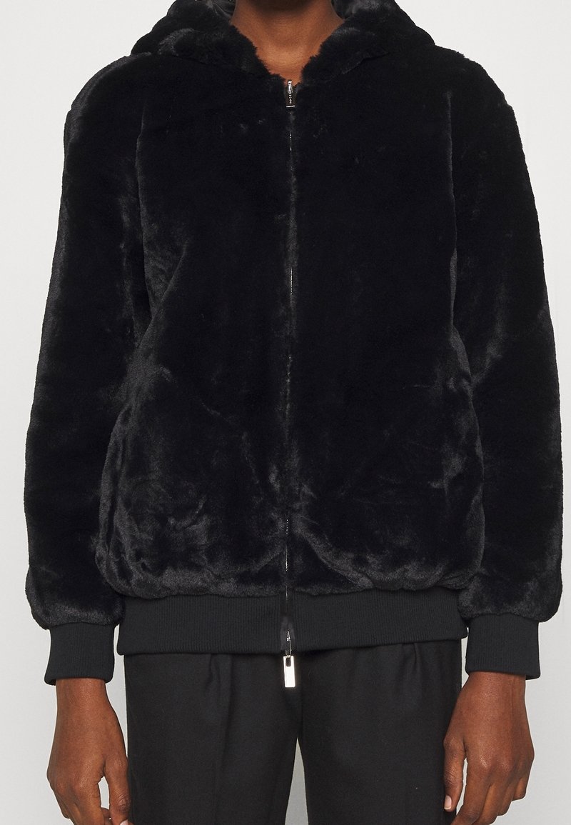 Zwarte pluche jack met rits en ribgebreide mouwen en onderrand, gedragen door een persoon, gecombineerd met zwarte broek, tegen een effen lichte achtergrond.