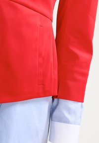 Veste rouge confectionnée avec une texture lisse, dotée d'une poche latérale et d'une coupe structurée. Poignets de chemise bleue avec des accents blancs visibles en dessous.