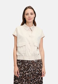 Beżowa, krótka kamizelka robocza z krótkimi rękawami, z guzikiem z przodu, dwiema kieszeniami na piersi oraz marszczonym dołem, połączona z plisowaną spódnicą w print w panterkę.