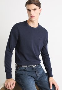 Dunkelblauer Strickpullover mit langen Ärmeln und Rundhalsausschnitt, versehen mit einem kleinen roten und weißen Logo auf der Brust, kombiniert mit dunklen Jeans.