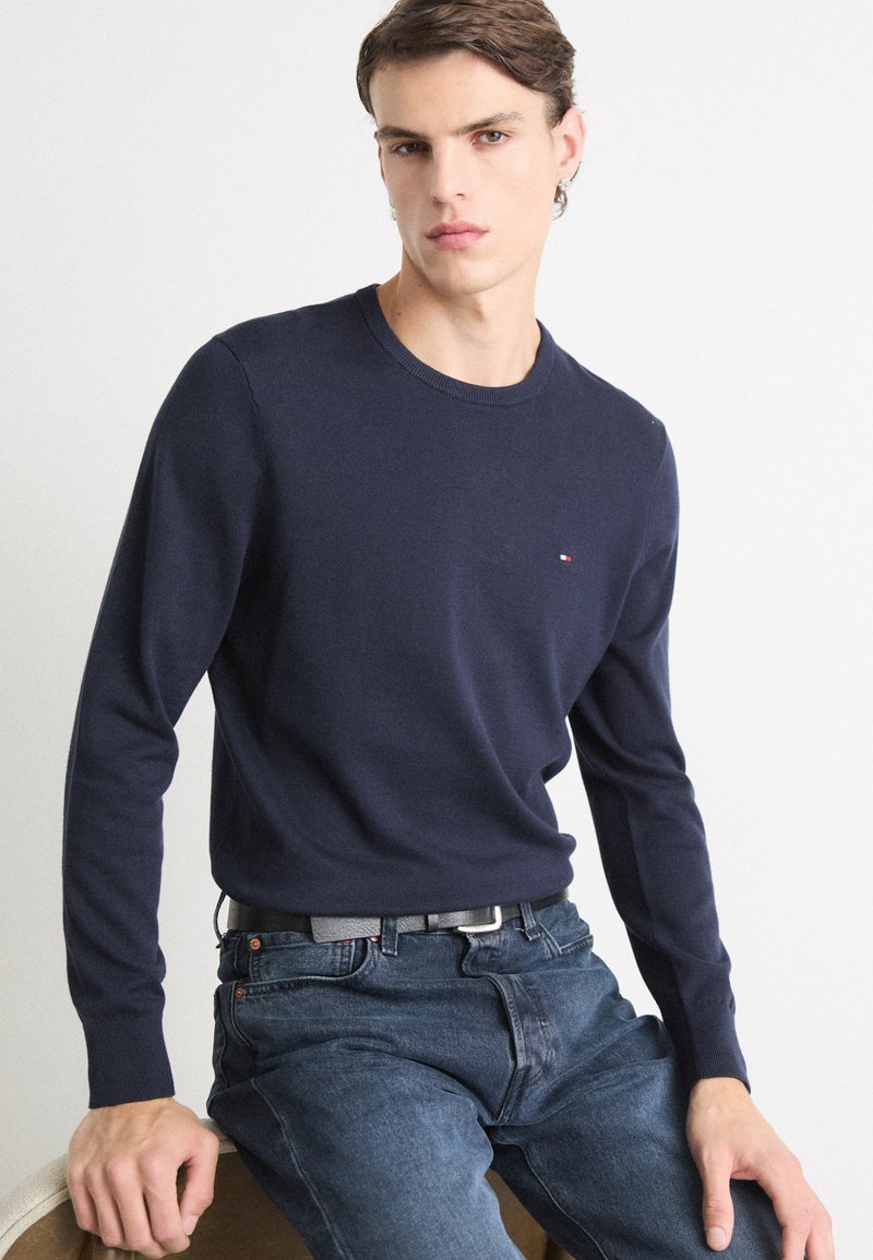 Dunkelblauer Strickpullover mit langen Ärmeln und Rundhalsausschnitt, versehen mit einem kleinen roten und weißen Logo auf der Brust, kombiniert mit dunklen Jeans.